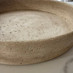 A close-up of a round, shallow tray or planter made of textured beige travertine stone, showing its natural speckled surface and thick rim.
