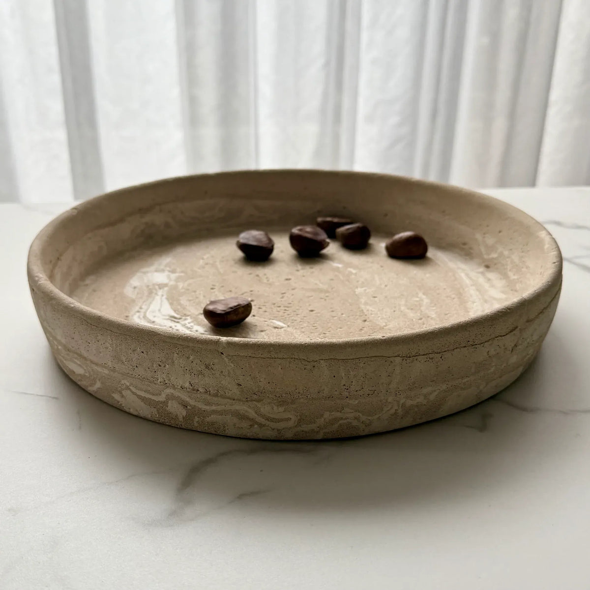 A round, shallow beige ceramic tray with a textured, hand-painted organic pattern, sitting on a white marble surface with a white curtain background. A few dark brown coffee beans are placed inside the tray.