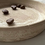 A close-up of a round, shallow ceramic plate or tray with a textured, speckled beige finish and a few dark brown coffee beans placed on top.
