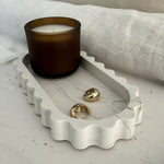 A white marble-look scalloped jewelry tray holding two gold rings, with an amber glass candle in the background on a light surface and draped fabric.
