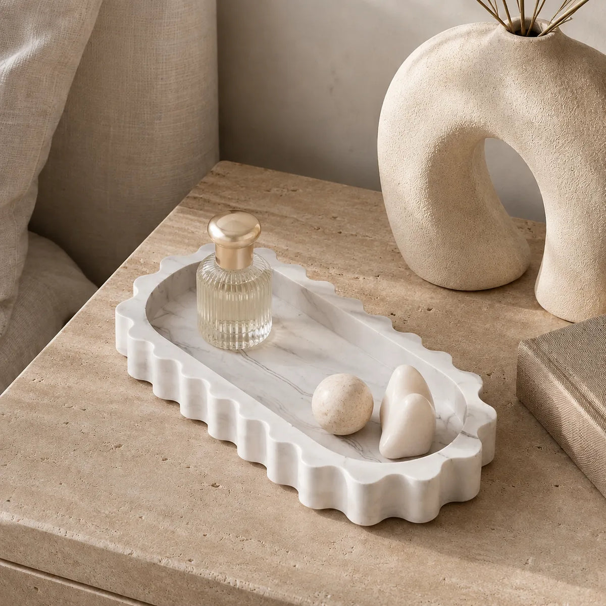 A white marble-look tray with a scalloped edge sits on a beige stone side table, styled with a small glass bottle, two stones, and a sculptural vase in a softly lit interior setting.