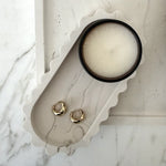 Two small gold hoop earrings displayed on a scalloped white marble tray next to a round white candle in a black container, set on a light marble surface.