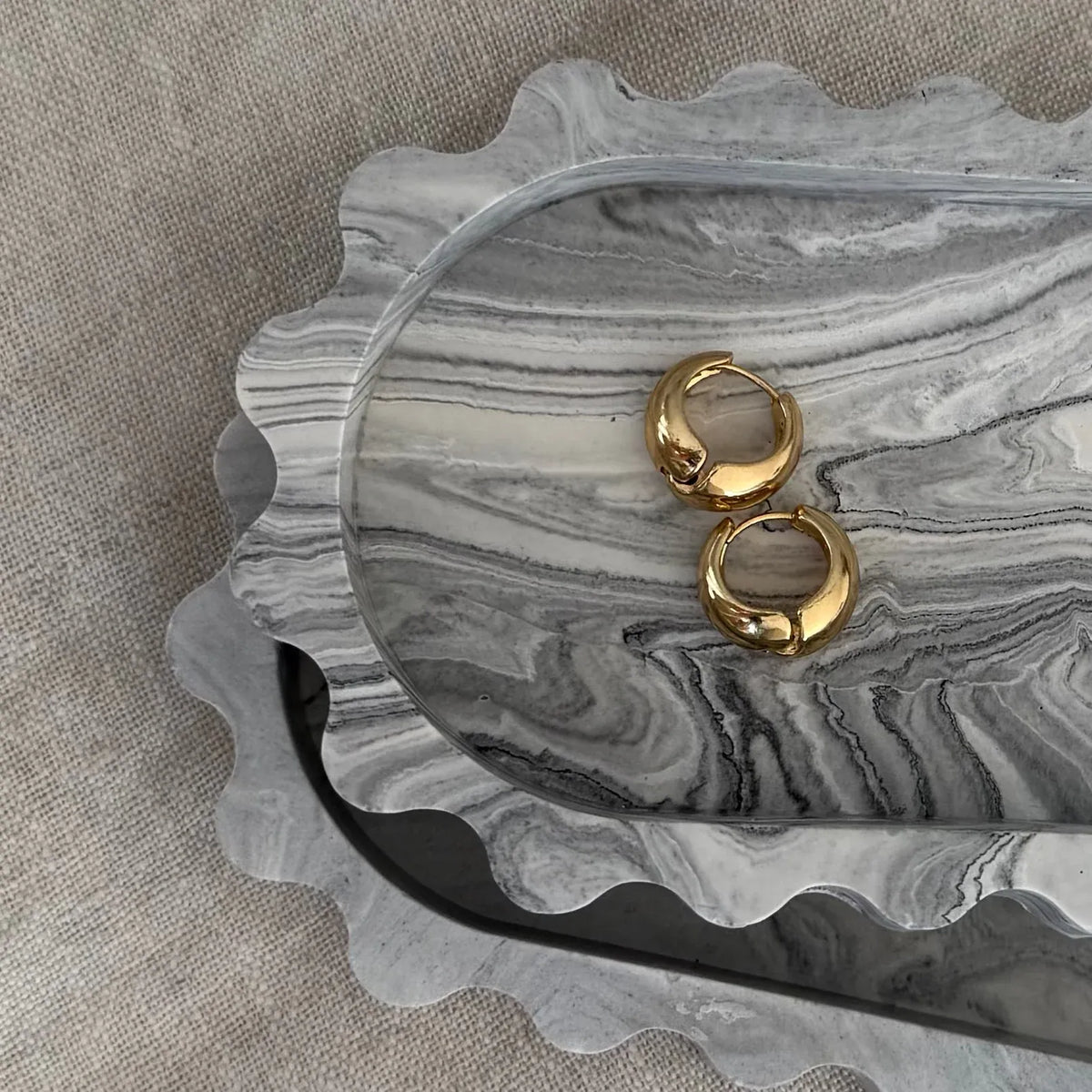 Two small gold hoop earrings displayed on a scalloped gray marbled tray over a beige fabric background.