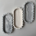 Three scalloped-edge rectangular trays with oval centers, arranged on a light gray background. Two trays have gray and white marble patterns, and the center tray is solid off-white with subtle marble veining.