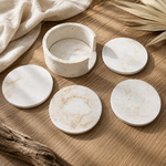 Five round off-white stone coasters with natural beige veining, including one open round cup, arranged on a wooden surface with neutral fabric and dried palm leaves.