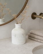 A ribbed white ceramic vase with dried baby's breath stems, styled on a bathroom counter next to folded towels and a round mirror.