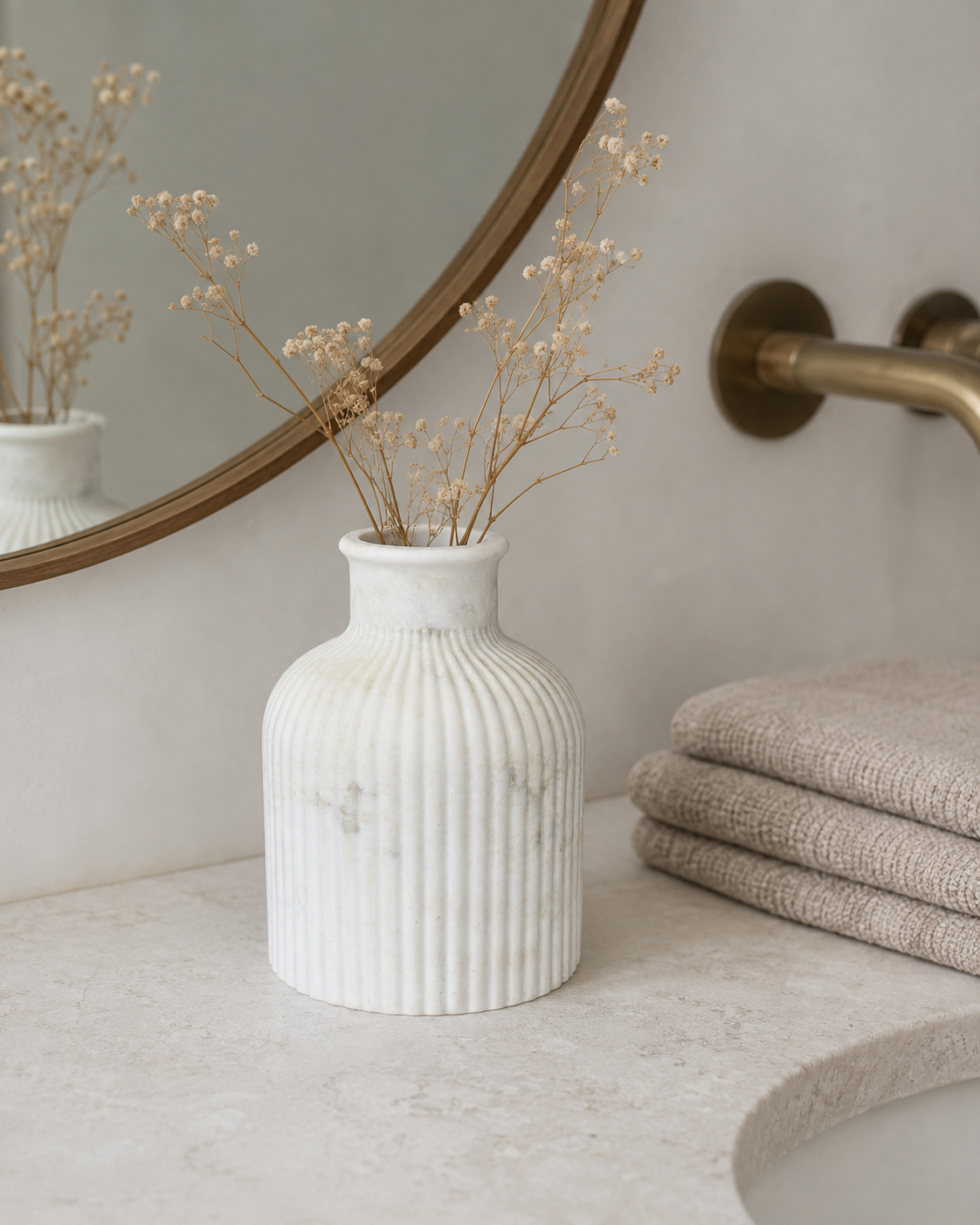 A ribbed white ceramic vase with dried baby's breath stems, styled on a bathroom counter next to folded towels and a round mirror.