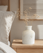 A white ribbed ceramic vase filled with dried baby's breath stems, styled on a light wood bedside table next to a bed with neutral-toned bedding.