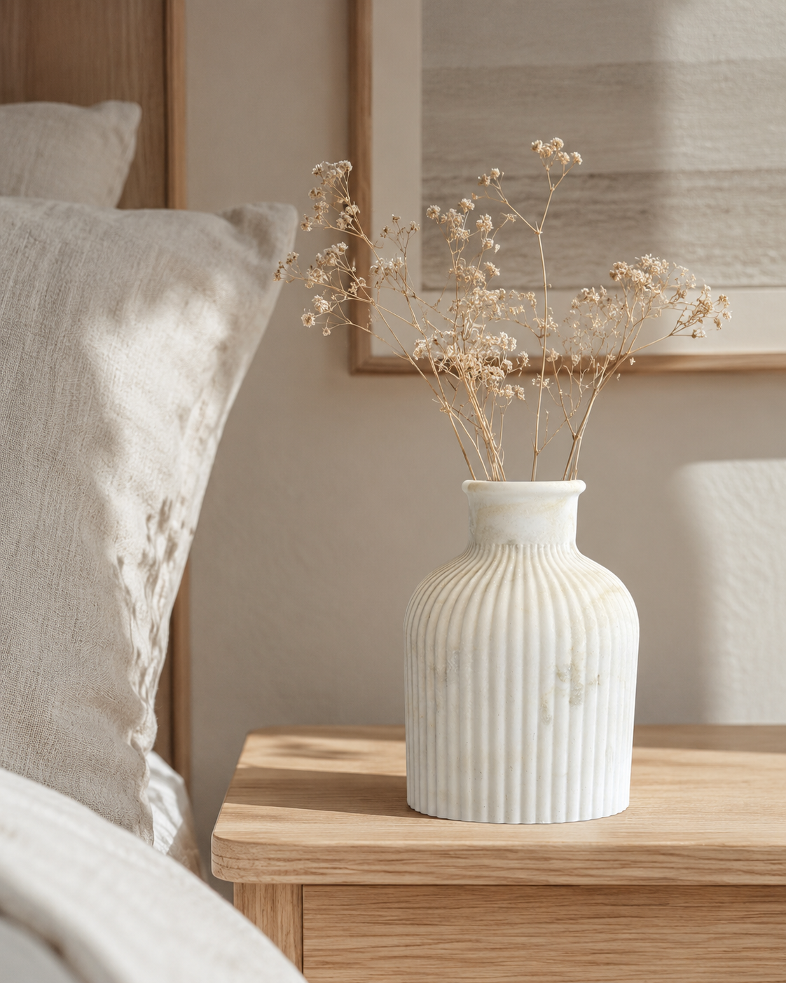 A white ribbed ceramic vase filled with dried baby's breath stems, styled on a light wood bedside table next to a bed with neutral-toned bedding.