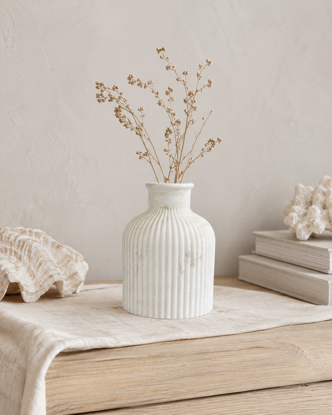 A white ribbed ceramic vase with dried botanical stems, styled on a light wood surface with a linen runner, a large clam shell, and stacked books against a textured beige wall.