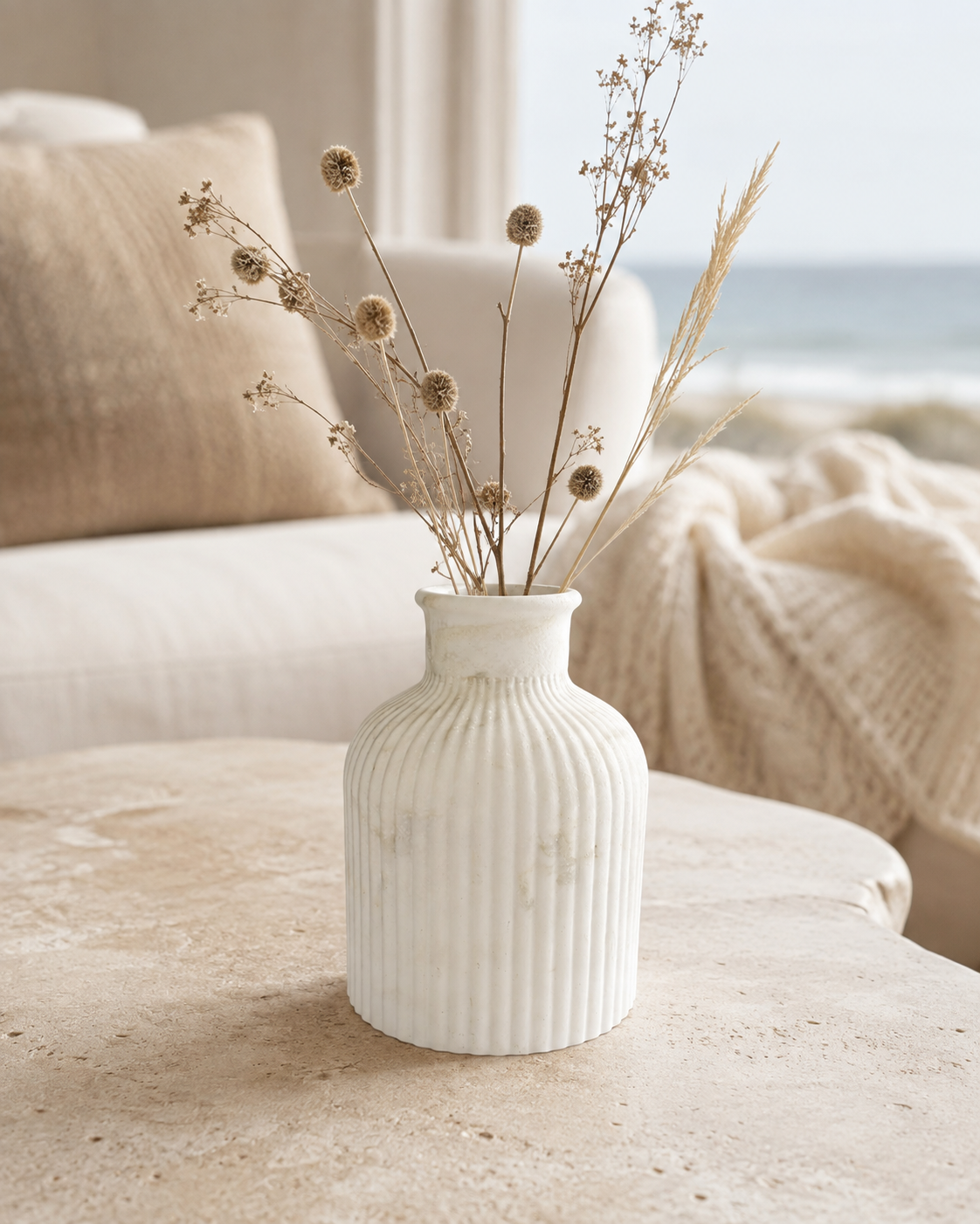 A white ribbed ceramic vase filled with dried grasses and small round flower heads, placed on a light stone table in a softly lit living room with a sofa, pillow, and blanket, and the ocean blurred in the background.
