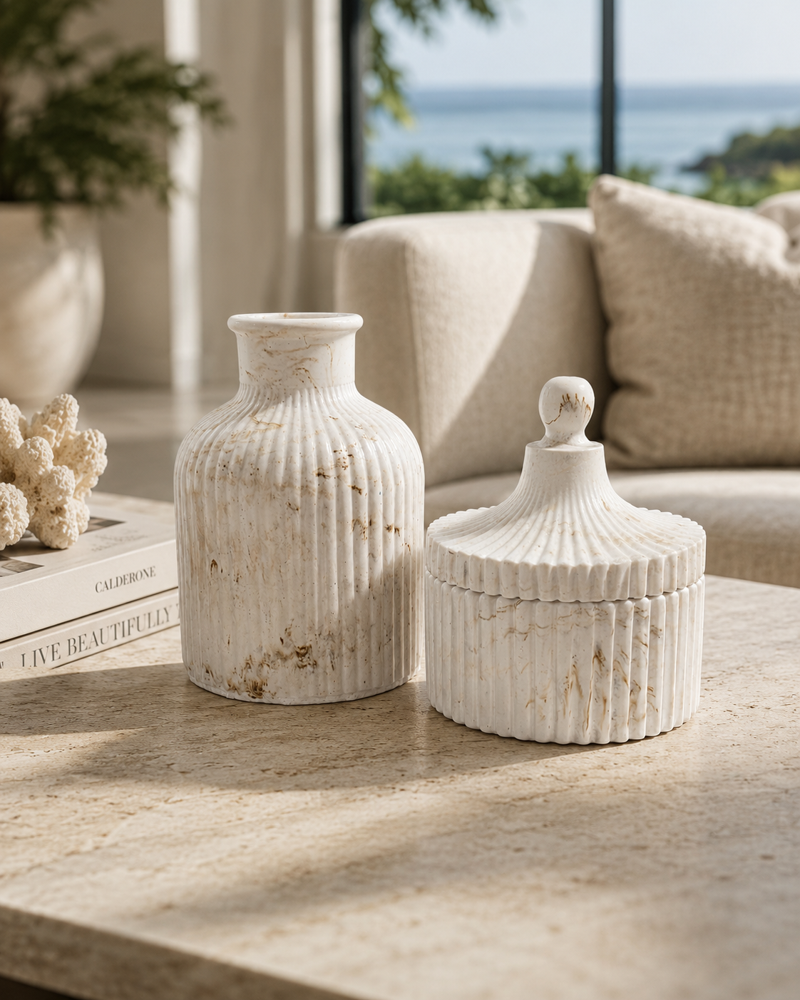 Two ribbed ceramic vases in an off-white marble finish, one standing as a vase and one as a lidded box with a knob, displayed on a stone table in a bright, modern living room.
