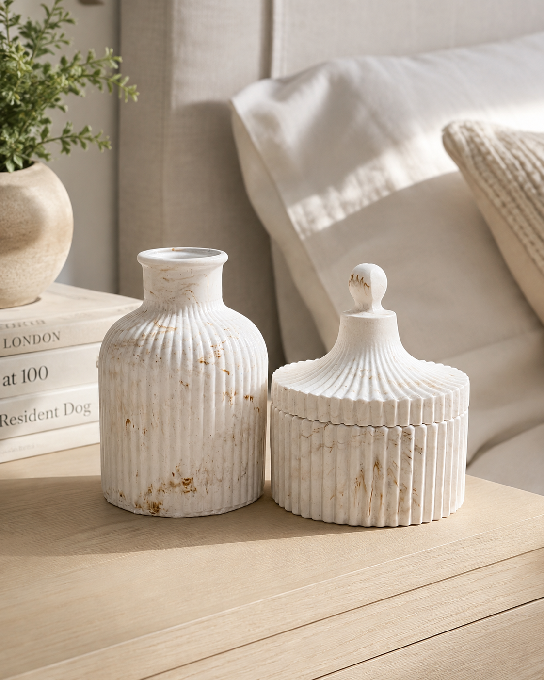 Two white ribbed decorative ceramic vases with a distressed gold finish, one standing as a vase and the other as a lidded box with a knob, displayed on a light wood table in a cozy bedroom setting.