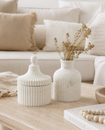 Two ribbed ceramic home decor pieces on a light wood table in a cozy living room setting: a white lidded jar with a rounded knob and a white ribbed vase holding dried beige stems, with cream pillows and a sofa in the background.