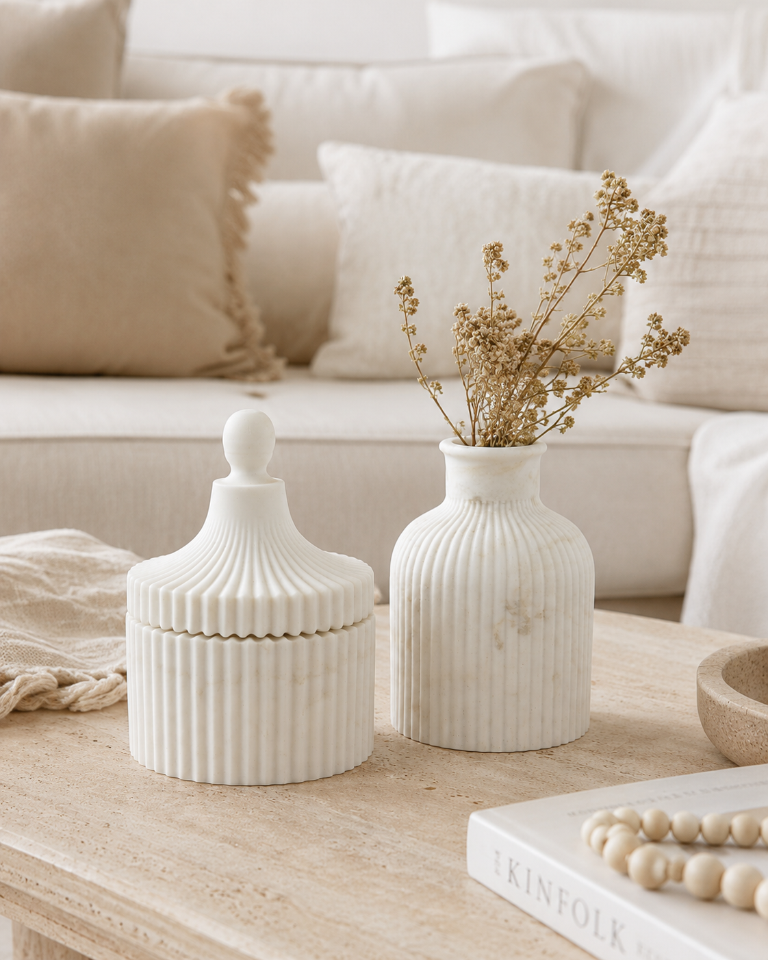 Two ribbed ceramic home decor pieces on a light wood table in a cozy living room setting: a white lidded jar with a rounded knob and a white ribbed vase holding dried beige stems, with cream pillows and a sofa in the background.