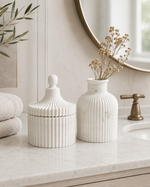 Two white ribbed ceramic containers, one lidded and one vase-shaped with dried flowers, styled on a marble bathroom counter.