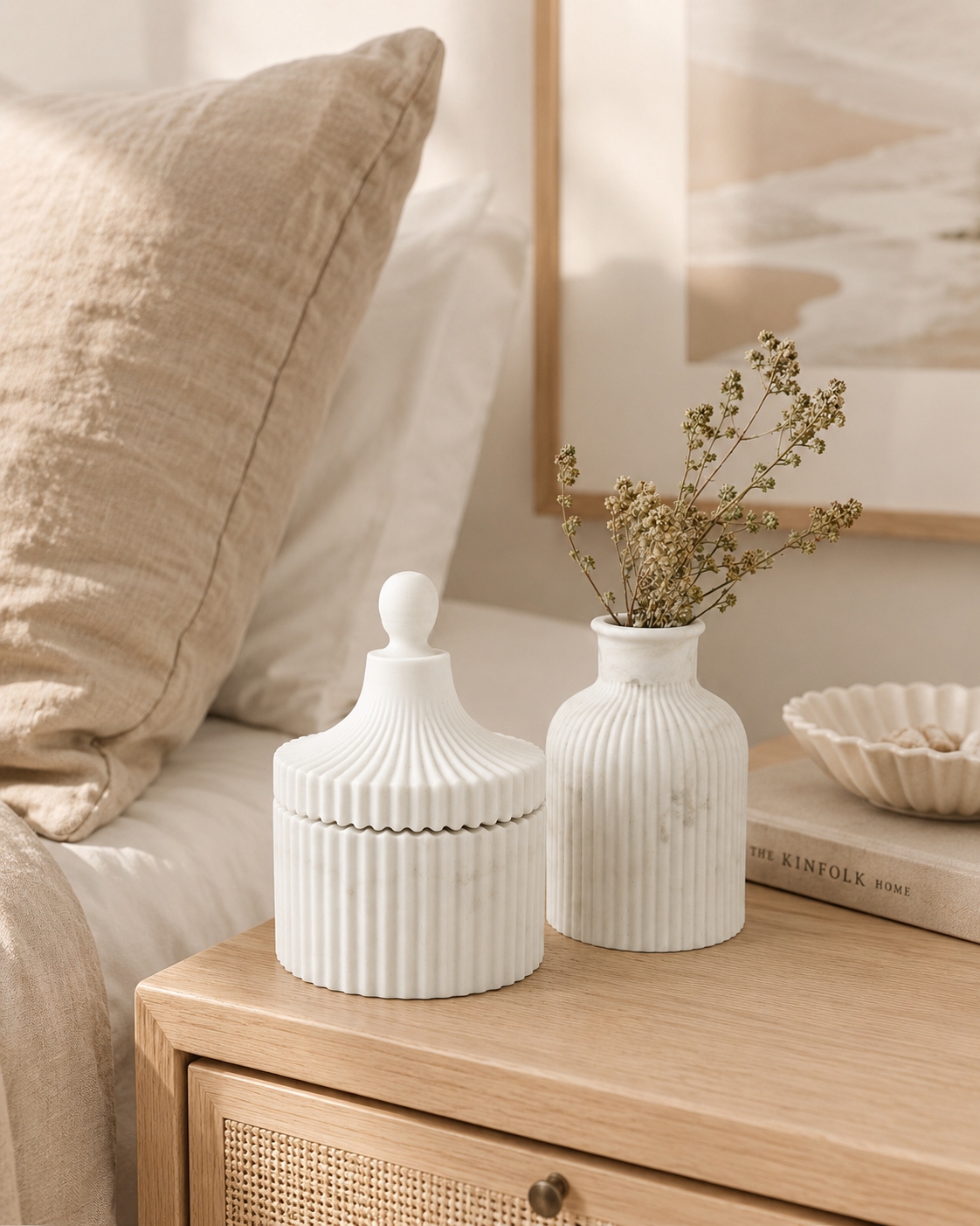 Two white ribbed ceramic decorative containers, one with a lid and one vase with dried stems, styled on a wooden bedside table in a warm, neutral bedroom setting.