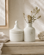Two white ribbed ceramic decorative containers on a light wood shelf: a lidded jar with a rounded knob top and a ribbed vase holding dried beige stems, set against a textured cream wall with soft natural shadows.