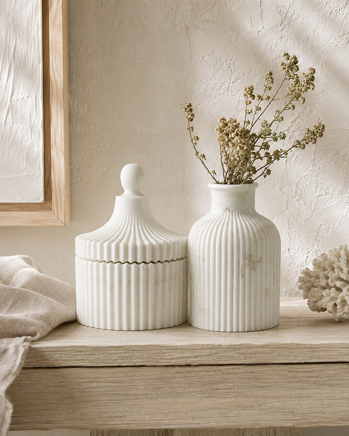 Two white ribbed ceramic decorative containers on a light wood shelf: a lidded jar with a rounded knob top and a ribbed vase holding dried beige stems, set against a textured cream wall with soft natural shadows.