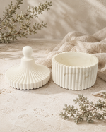A white ceramic jewelry box with a matching lid, both featuring a fluted ribbed texture and a spherical finial on top, styled on a textured beige surface with linen and dried botanicals.