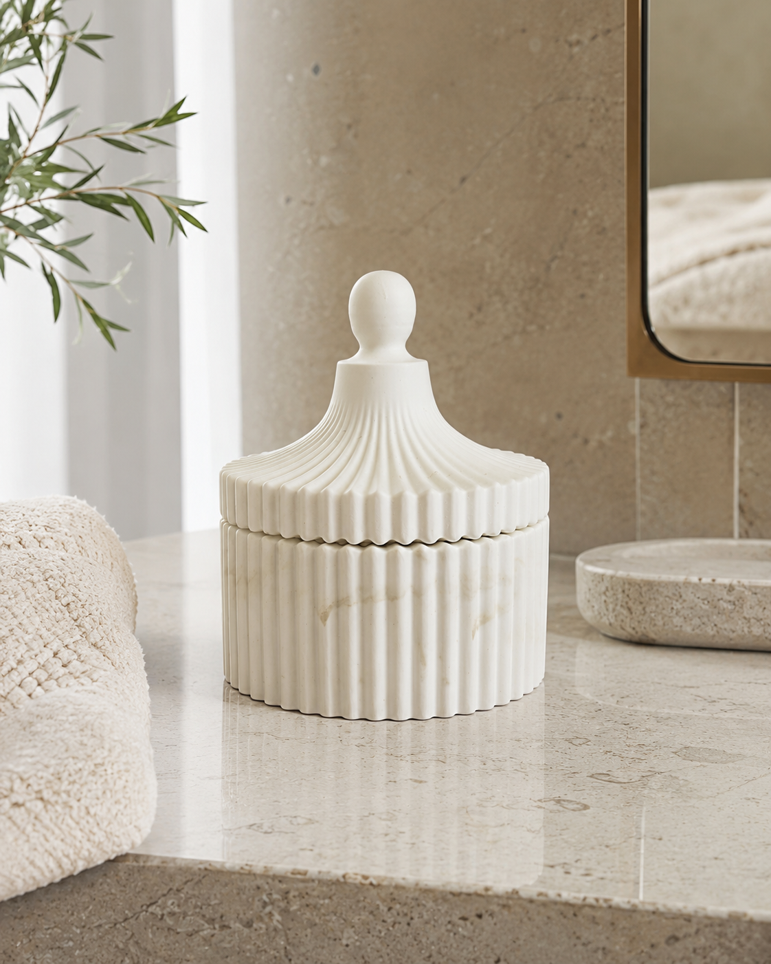 A ribbed white ceramic lidded container with a rounded knob top, styled on a beige marble bathroom counter next to a folded cream towel and a mirror.