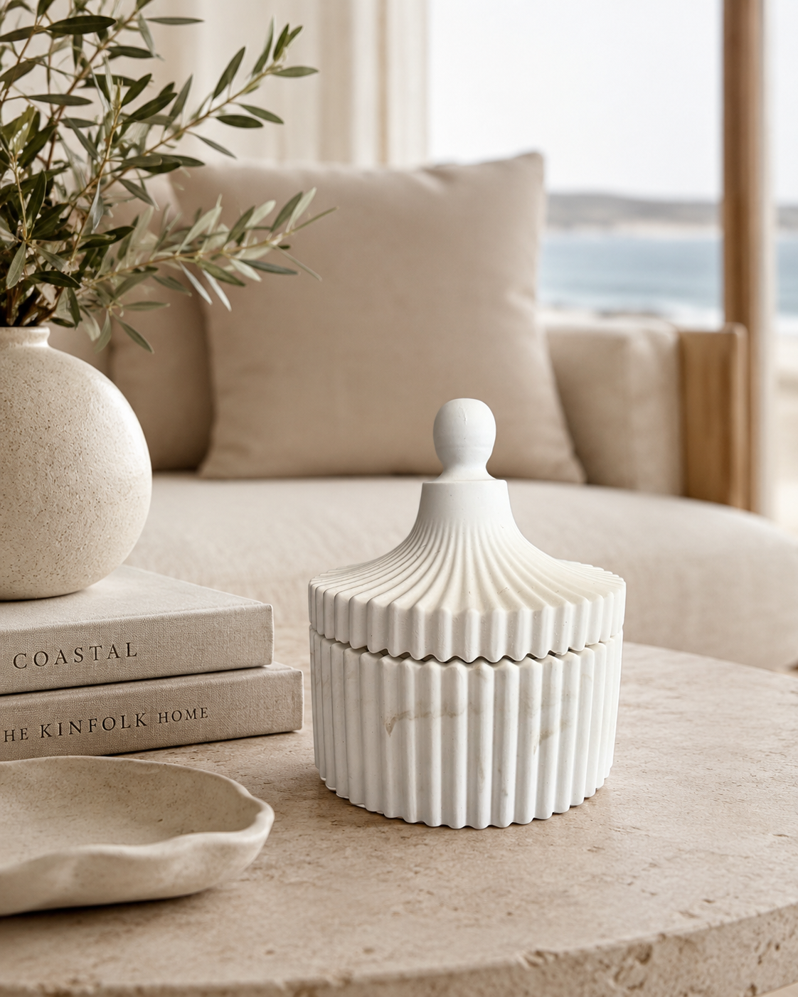 A ribbed white ceramic lidded box with a rounded knob top, styled on a stone table next to a vase and books in a bright, modern living room setting.