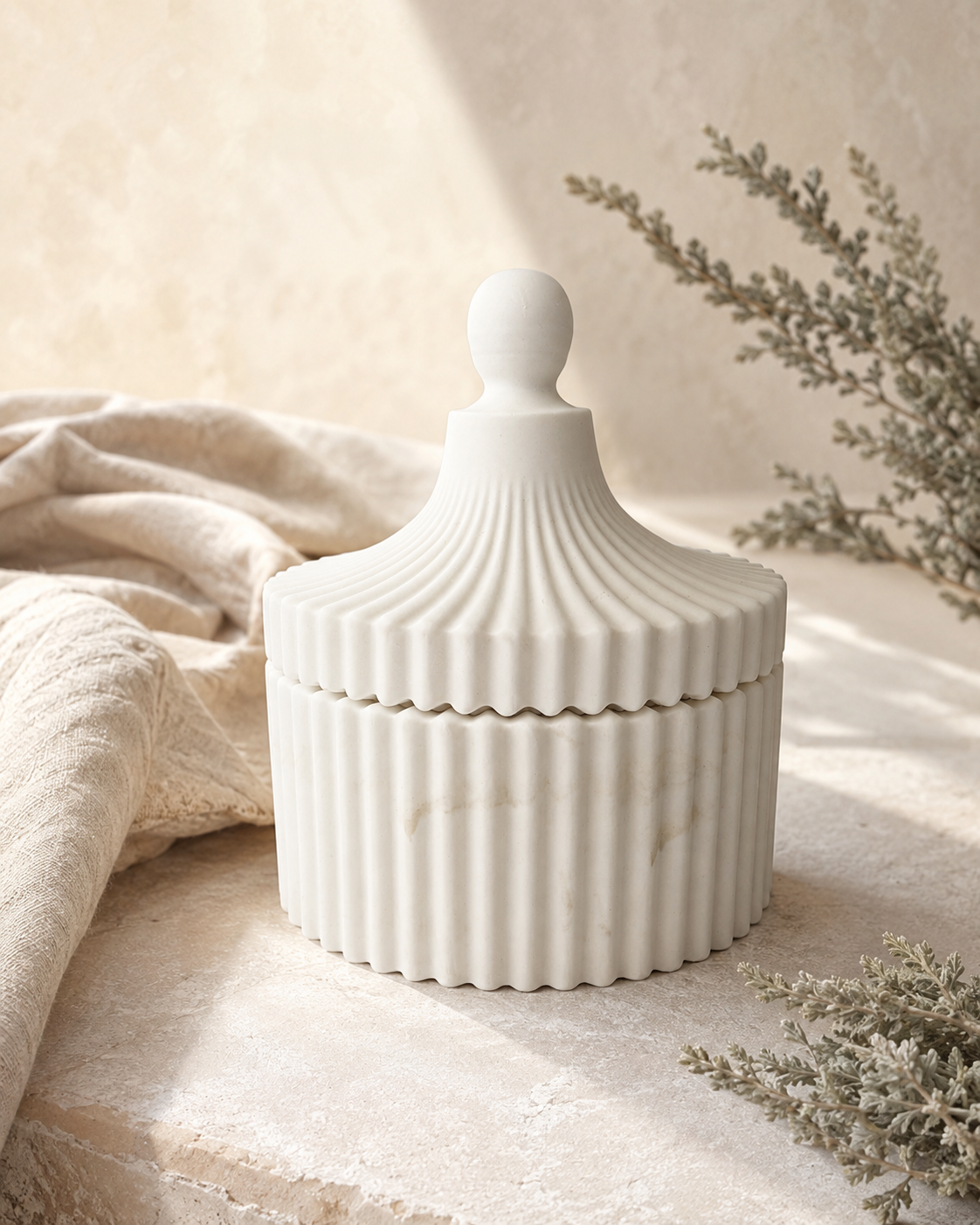 A white ceramic lidded jar with a fluted ribbed design and a round knob, styled on a stone surface with a beige linen cloth and dried botanicals.