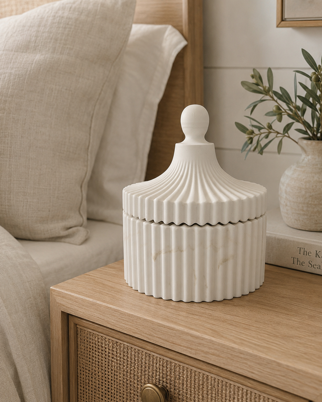 A ribbed ceramic lidded jar in warm off-white sits on a light wood bedside table next to a bed with beige bedding and a small vase with greenery.