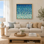 A framed abstract painting with textured blue, teal, and white brushstrokes hangs on a beige wall above a light cream sofa in a bright, modern living room.