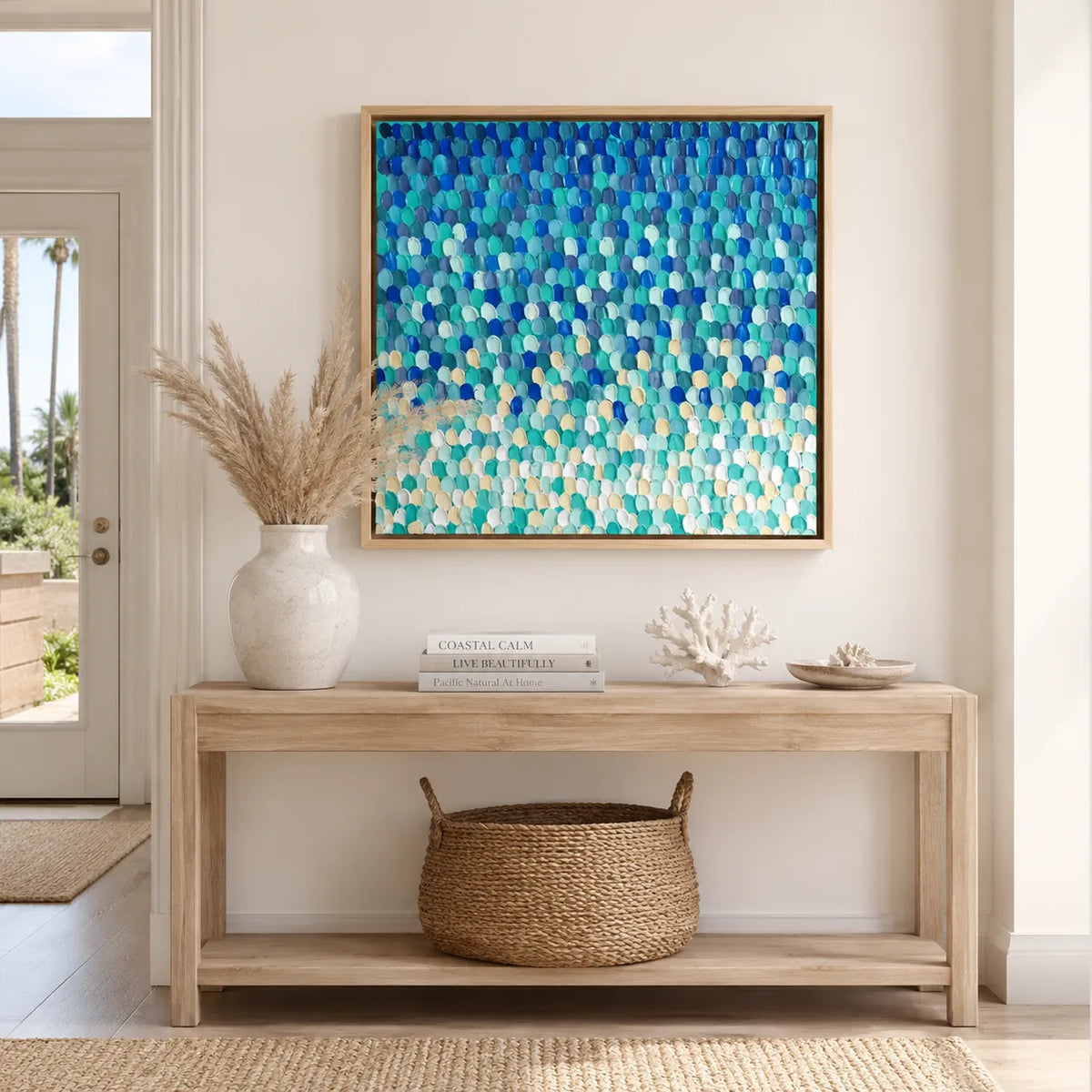 A square framed wall art print featuring an abstract textured pattern of overlapping scallop-like shapes in shades of blue, teal, and aqua, hanging on a beige wall above a light wood console table with decorative items.