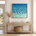 A square framed wall art print featuring an abstract textured pattern of overlapping scallop-like shapes in shades of blue, teal, and aqua, hanging on a beige wall above a light wood console table with decorative items.