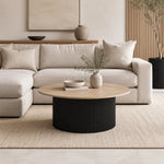 A modern living room scene featuring a light beige sectional sofa with matching pillows, a round coffee table with a black ribbed base and light wood top, and neutral decor on a woven rug.