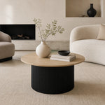 A round coffee table in a modern living room setting, featuring a light natural wood top and a ribbed black cylindrical base. The table is styled with a cream ceramic vase containing green branches, a stack of two books, and a small black bowl.