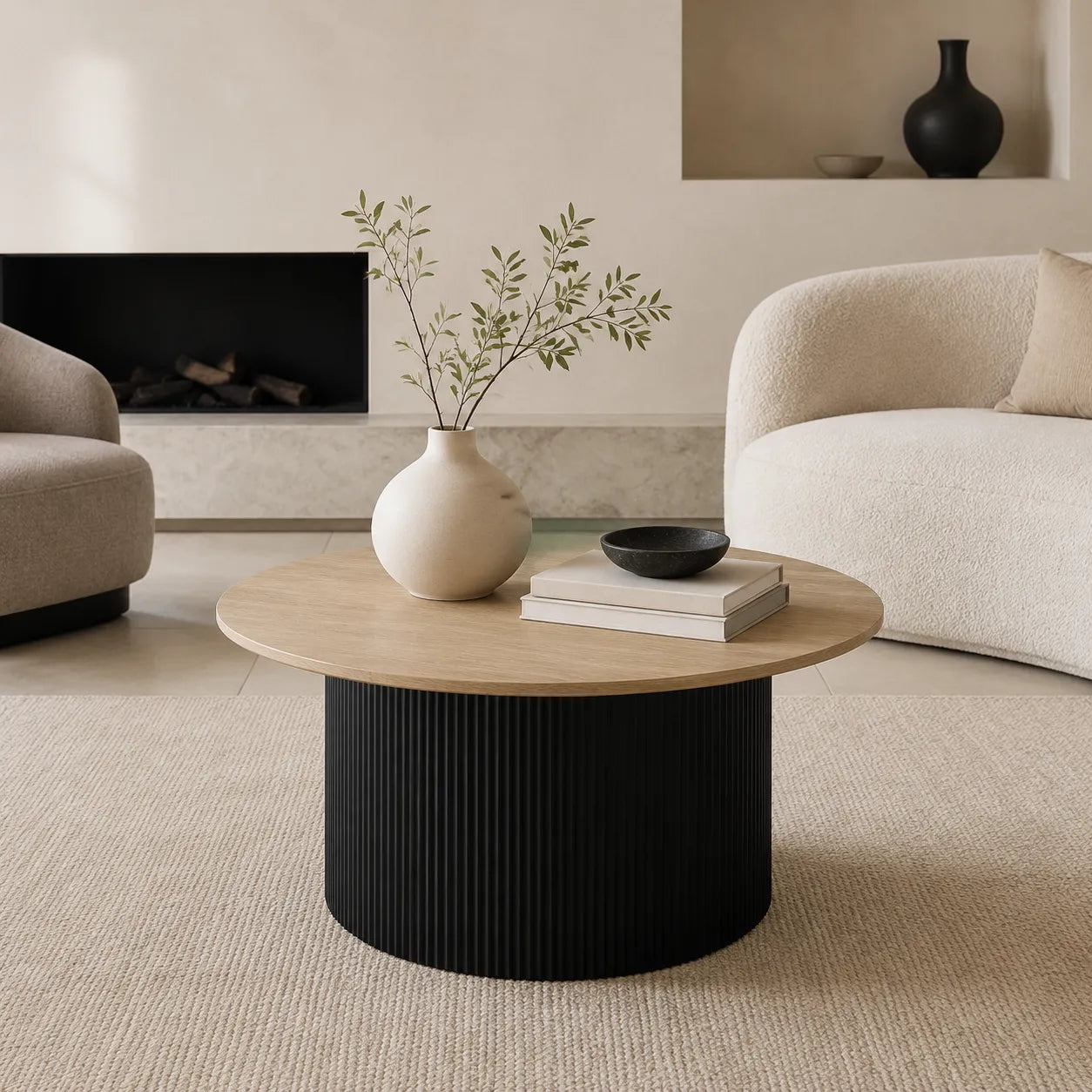 A round coffee table in a modern living room setting, featuring a light natural wood top and a ribbed black cylindrical base. The table is styled with a cream ceramic vase containing green branches, a stack of two books, and a small black bowl.