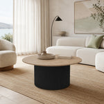 A modern round coffee table with a light wood top and a dark ribbed cylindrical pedestal base, styled in a bright, contemporary living room next to a white sofa and beige rug.