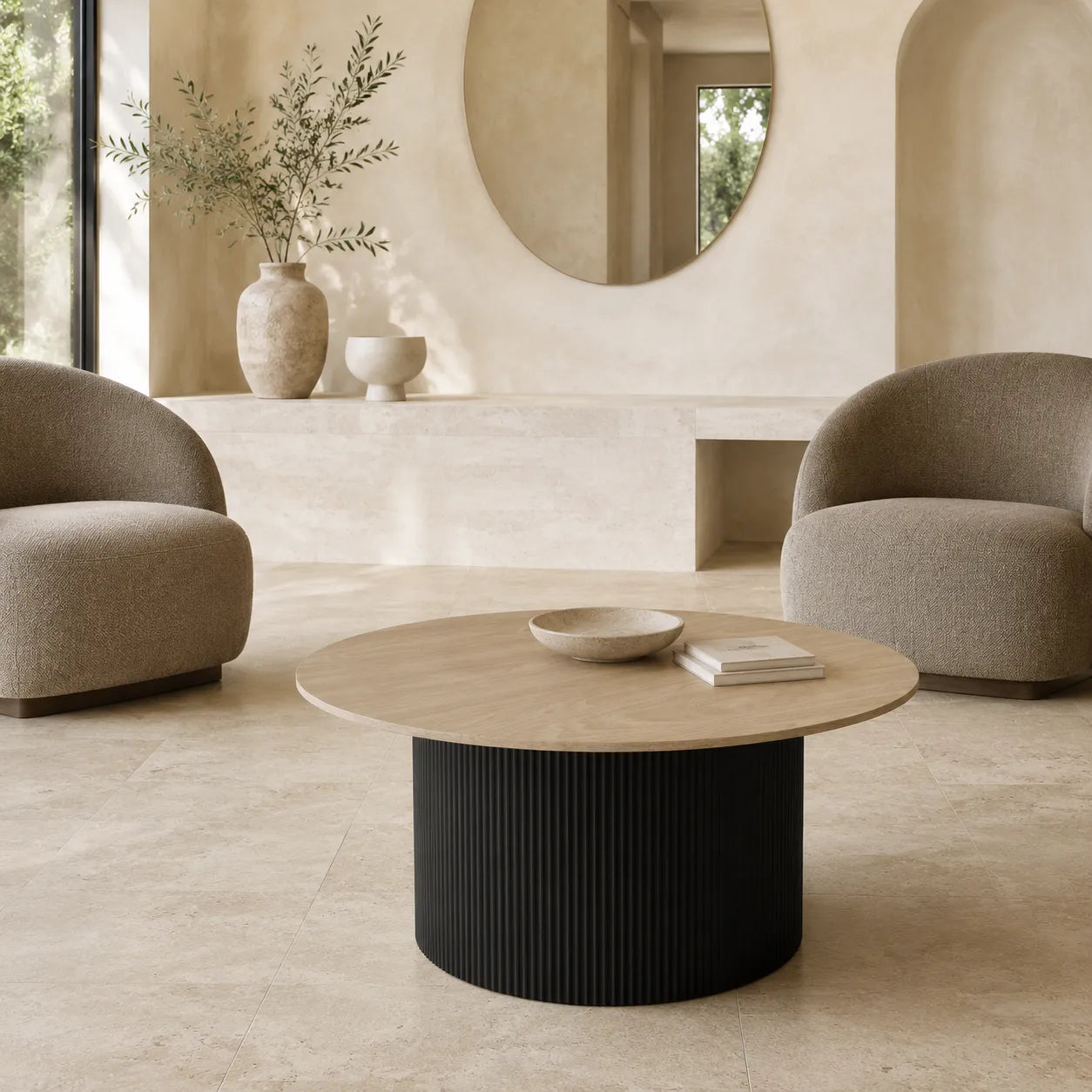 A modern round coffee table with a light wood top and a ribbed black pedestal base, styled in a minimalist, warm-toned living room with two beige armchairs.