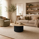 A modern living room scene featuring a beige fabric sofa, a matching armchair, a round coffee table with a light wood top and ribbed black base, set on a large textured cream rug. The room has warm neutral tones, built-in wooden shelves, and a tall olive tree near a window.