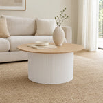 A modern round coffee table with a light wood top and a ribbed white cylindrical base, styled in a bright living room with a sofa, rug, and a vase on top.