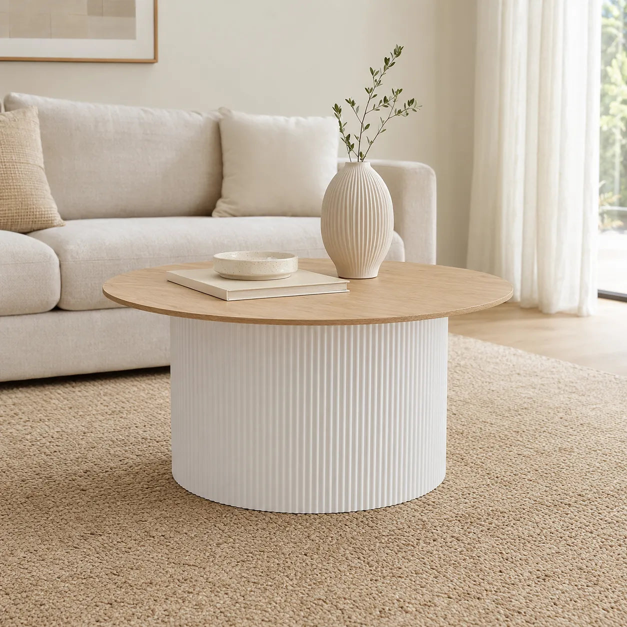 A modern round coffee table with a light wood top and a ribbed white cylindrical base, styled in a bright living room with a sofa, rug, and a vase on top.