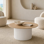 A round coffee table with a light natural wood top and a ribbed white cylindrical base, styled in a minimalist, warm-toned living room with two textured armchairs.
