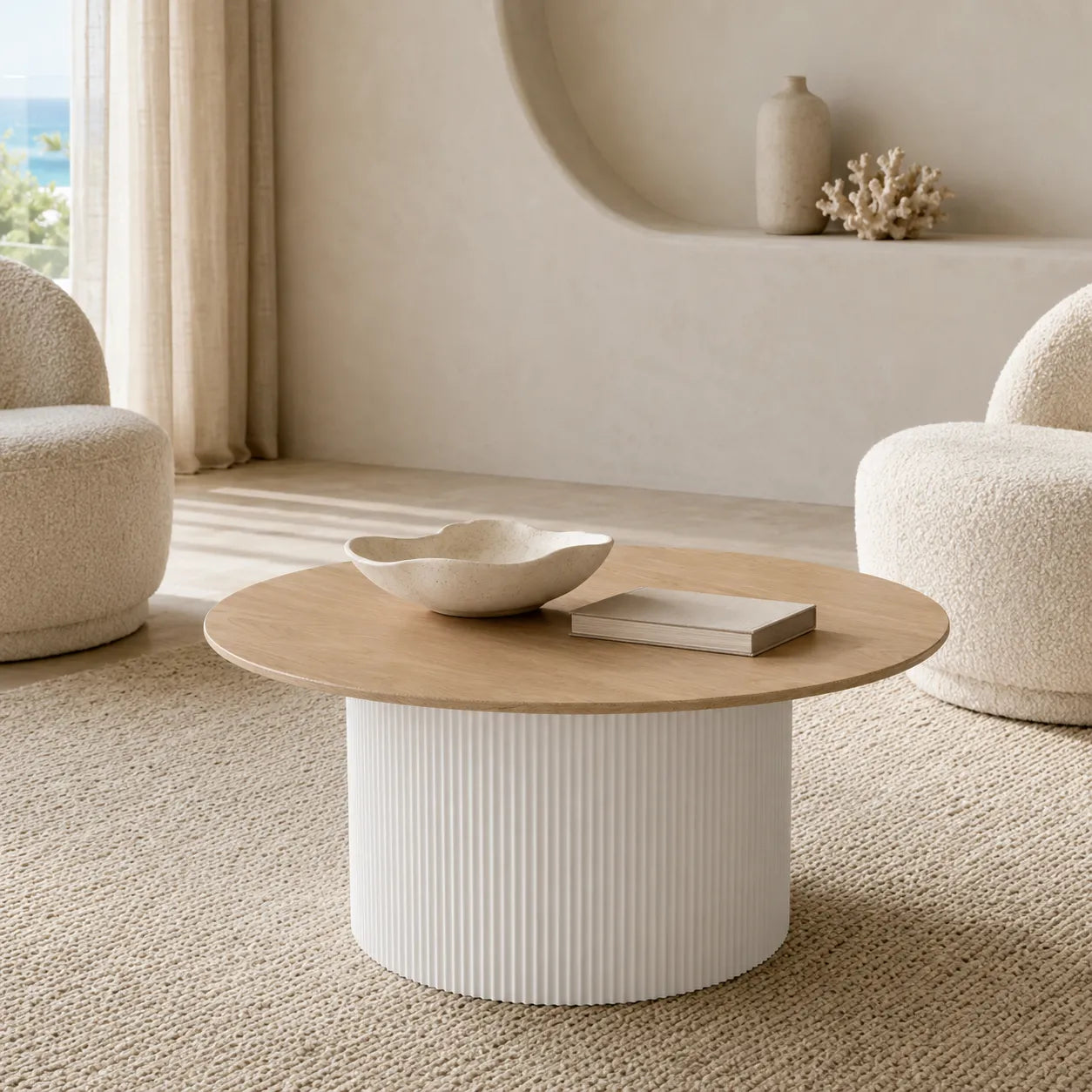 A round coffee table with a light natural wood top and a ribbed white cylindrical base, styled in a minimalist, warm-toned living room with two textured armchairs.