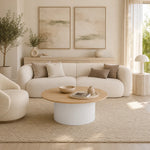 A modern living room featuring a white curved sofa with neutral throw pillows, a round wooden coffee table on a textured beige rug, and two large abstract art prints on the wall.