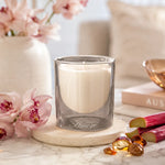 A white candle in a clear glass jar sits on a round stone coaster on a marble surface, styled with pink flowers, a vase, books, and rhubarb in a bright indoor setting.