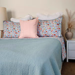 Bed with floral patterned pillows and a light blue comforter, nightstand with decorative vase.