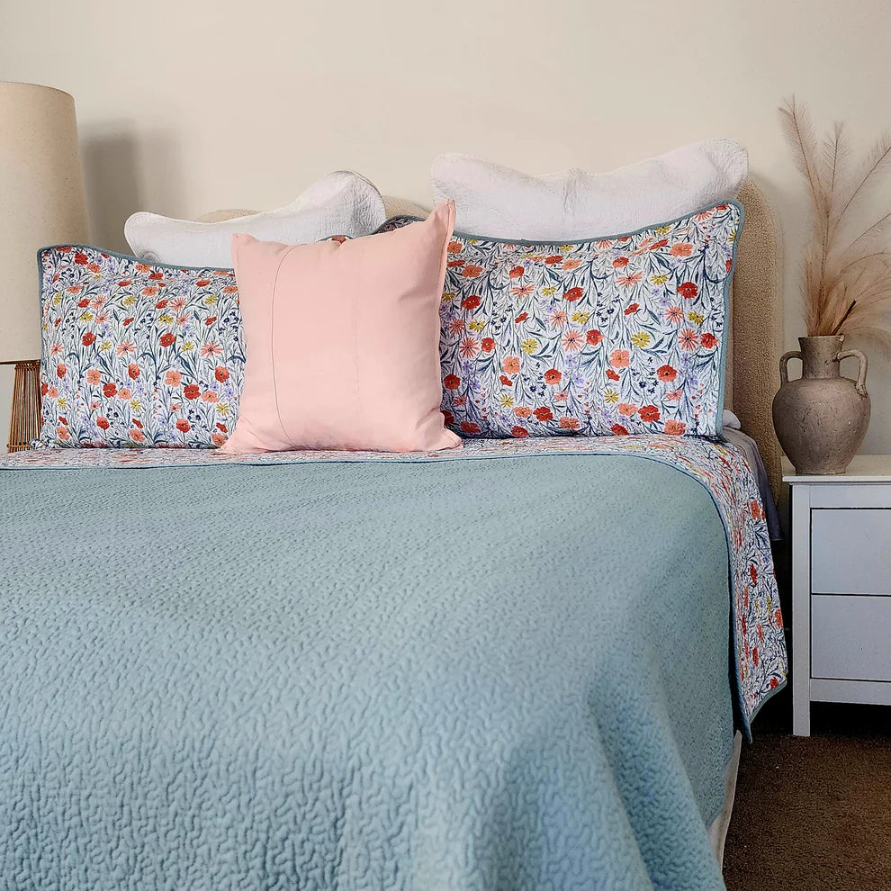 Bed with floral patterned pillows and a light blue comforter, nightstand with decorative vase.