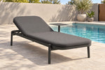 A modern outdoor chaise lounge chair with dark gray cushions and a black metal frame, positioned next to a swimming pool on a light stone patio.
