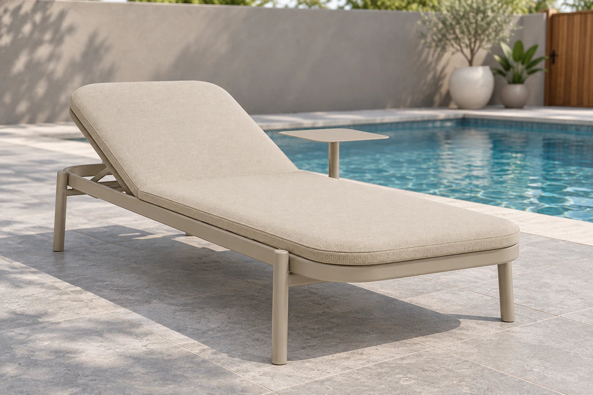 A modern outdoor chaise lounge with a light beige cushioned seat and a matching metal frame, positioned next to a swimming pool on a stone patio.
