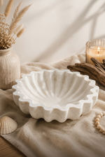 A scalloped white ceramic bowl with a ribbed, shell-like edge, styled on a beige linen cloth with dried grass in a vase, driftwood, and a lit candle in the background.