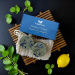Two soap bars with spearmint, lemon, and peppermint on a natural fiber mat with a blue box labeled 'Calm & Cozy' on a dark surface.