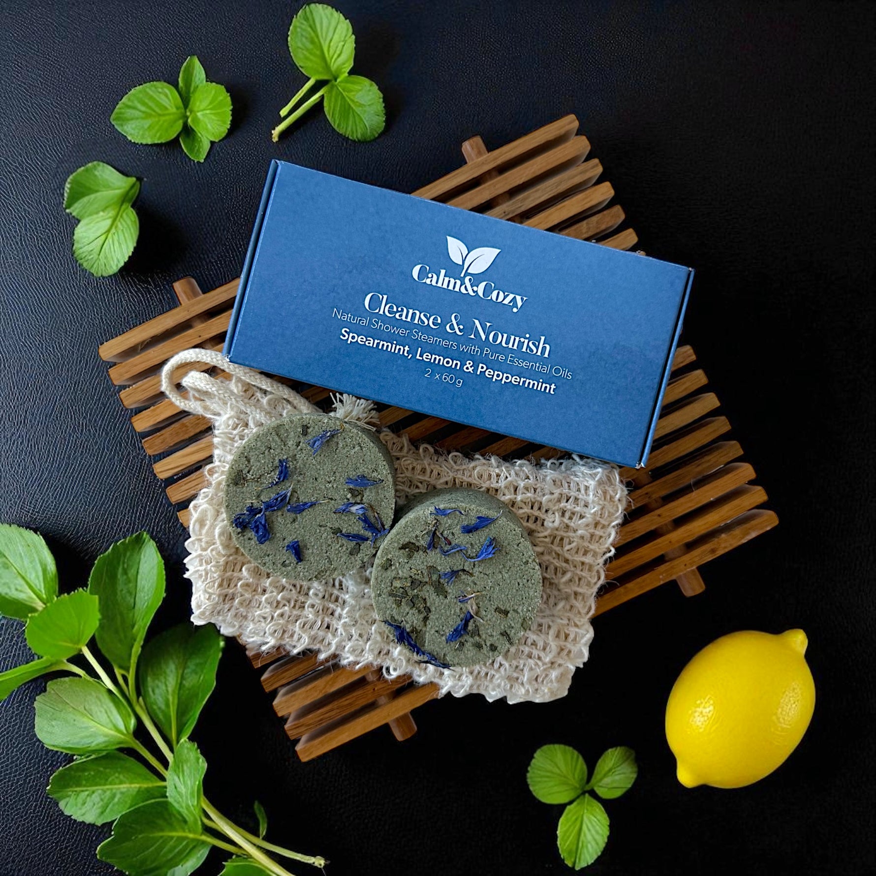 Two soap bars with spearmint, lemon, and peppermint on a natural fiber mat with a blue box labeled 'Calm & Cozy' on a dark surface.